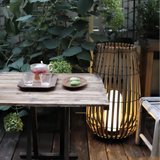 Tall woven rattan outdoor floor lantern beside a rustic wooden garden dining table