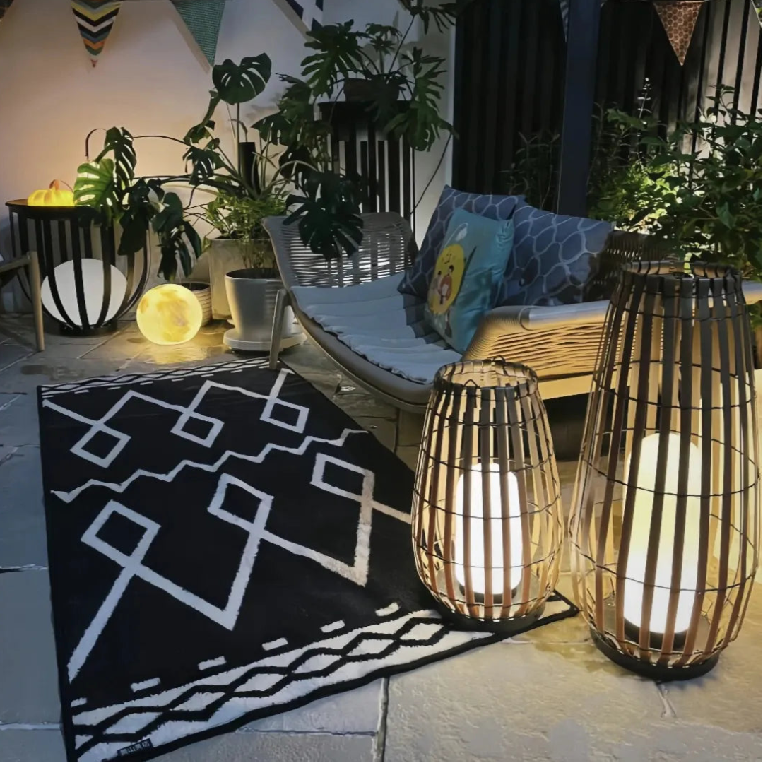Two glowing rattan floor lanterns decorating a cosy outdoor seating area with a patterned rug