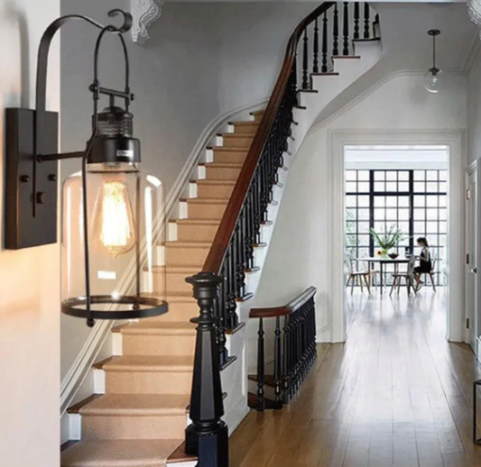 Black metal and glass wall lamp lighting a hallway beside a staircase, adding warm ambient light and enhancing a modern home interior.