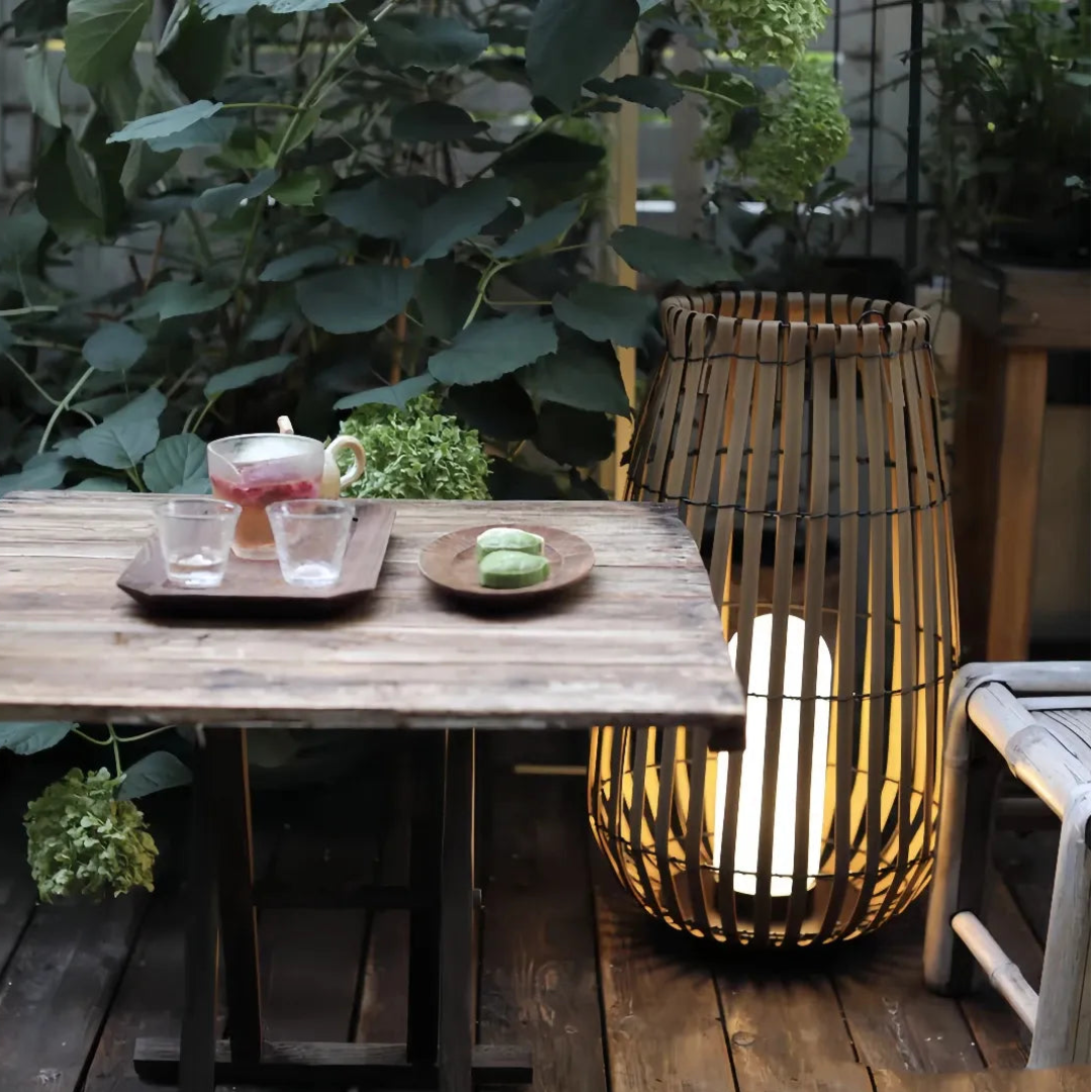 Tall woven rattan outdoor floor lantern beside a rustic wooden garden dining table