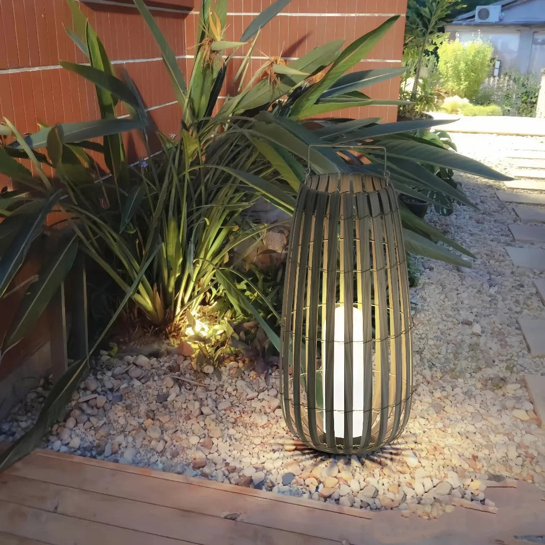Tall bohemian rattan garden lantern with warm LED light among plants and decorative gravel